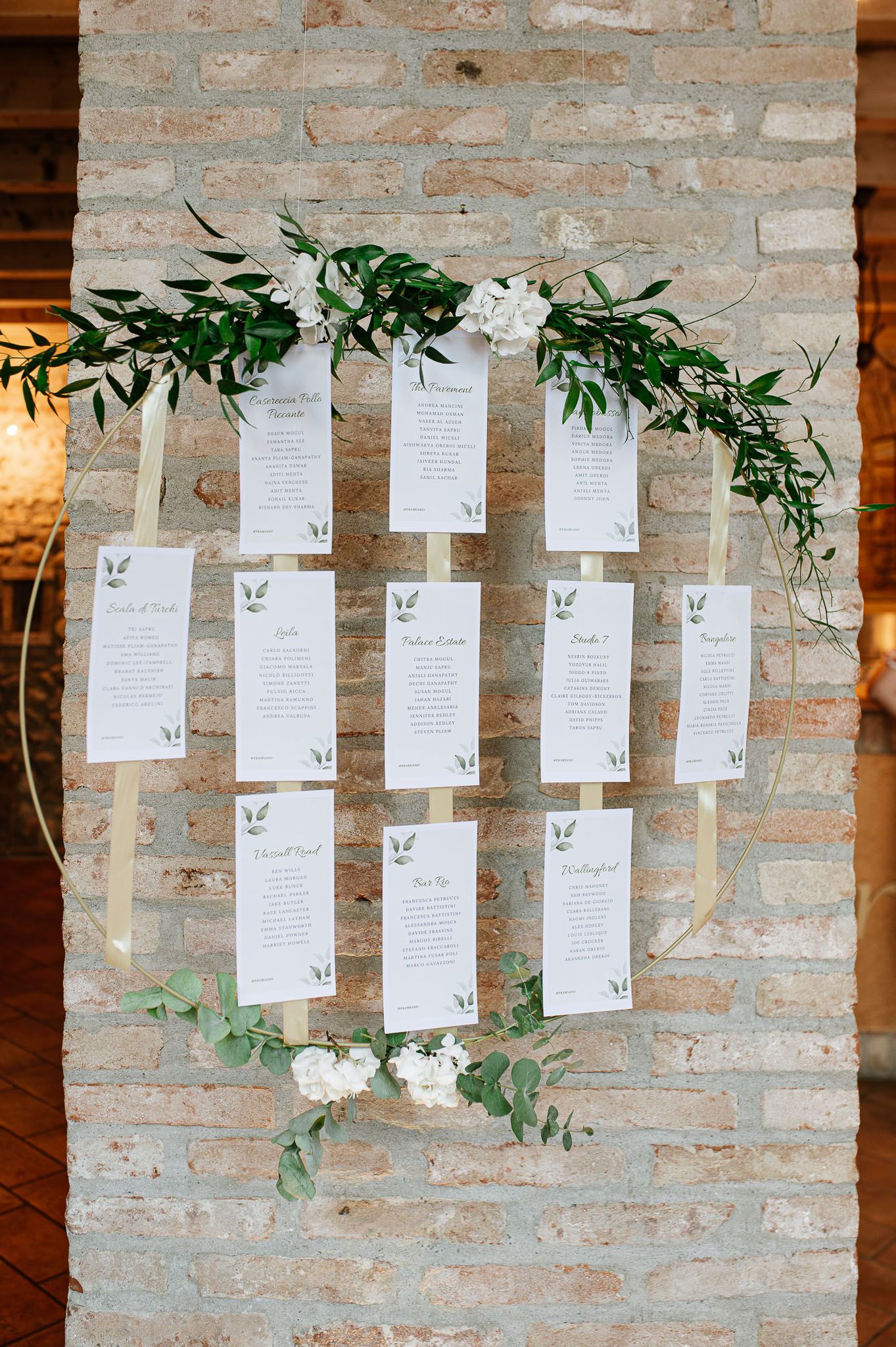 Wedding seating chart display with white cards and greenery decorations on a brick wall.