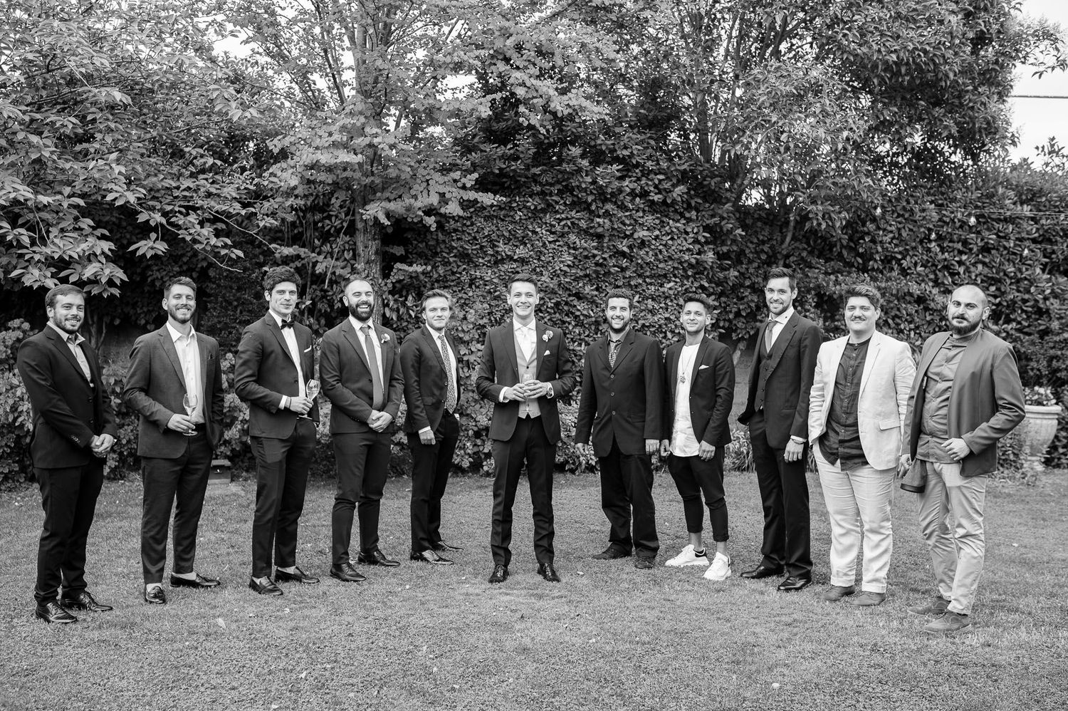 Black and white photo of groomsmen lined up outdoors in formal attire.