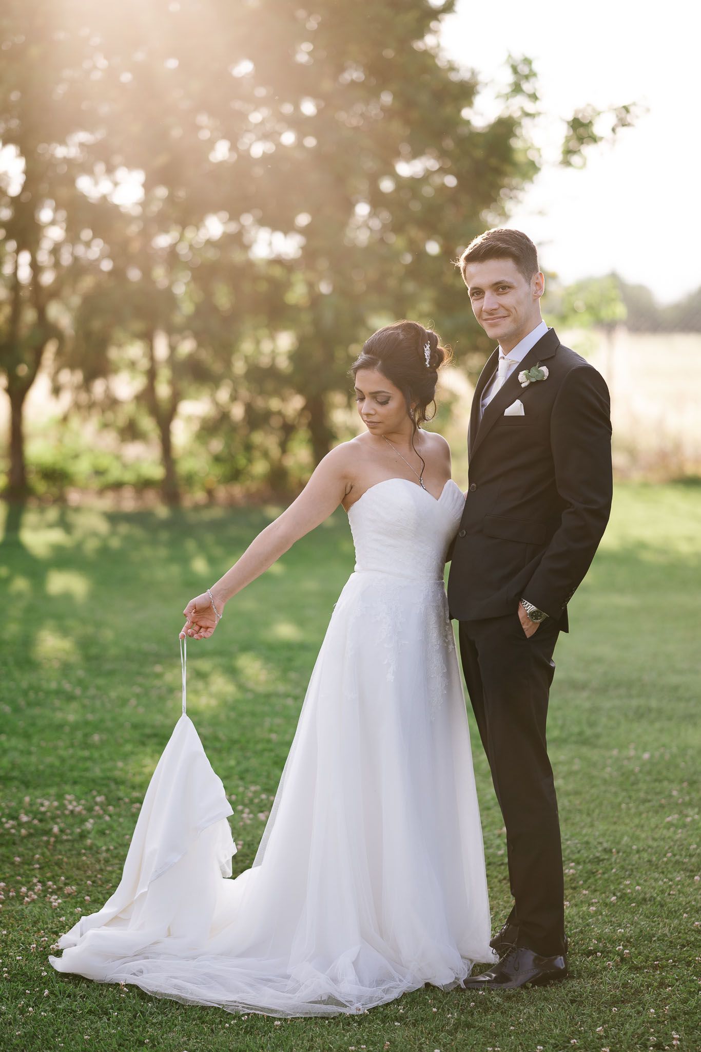 Newlywed couple poses outdoors in scenic setting with flowing white wedding dress and classic black tuxedo.