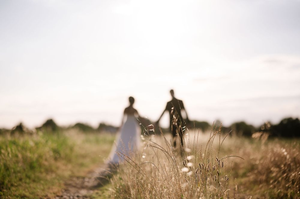 Silhouette of wedding couple holding hands while walking through tall grass at sunset.