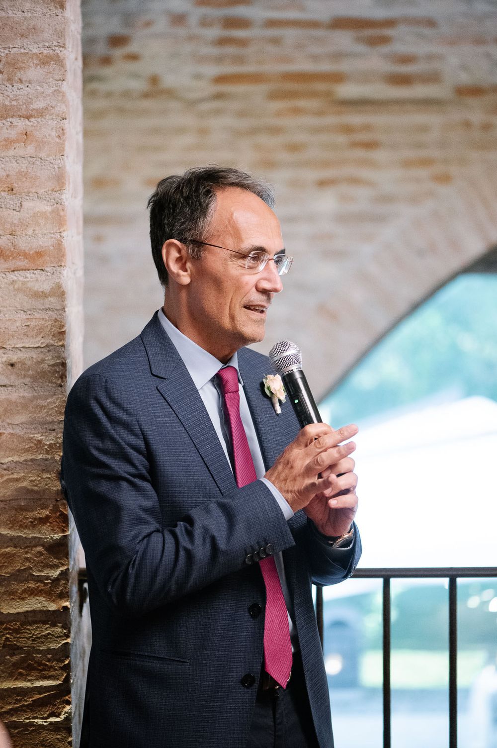 Speaker in navy suit delivers wedding speech at elegant brick venue with arched windows.