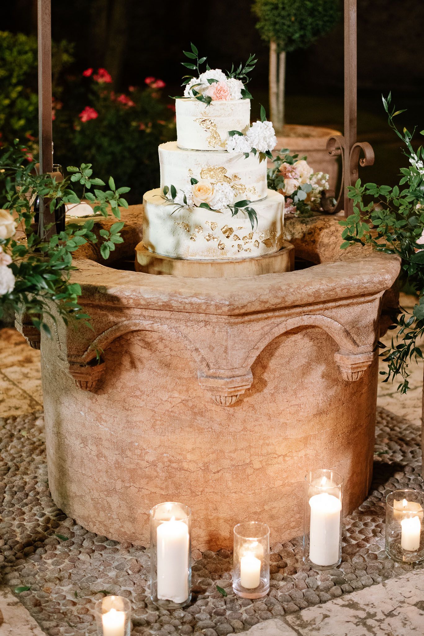 Elegant white wedding cake decorated with flowers sits atop stone pedestal surrounded by glowing candles.