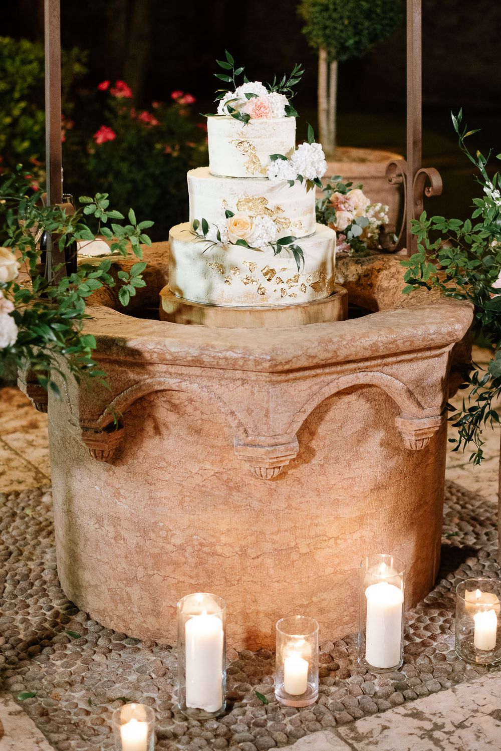 Elegant white wedding cake decorated with flowers sits atop stone pedestal surrounded by glowing candles.