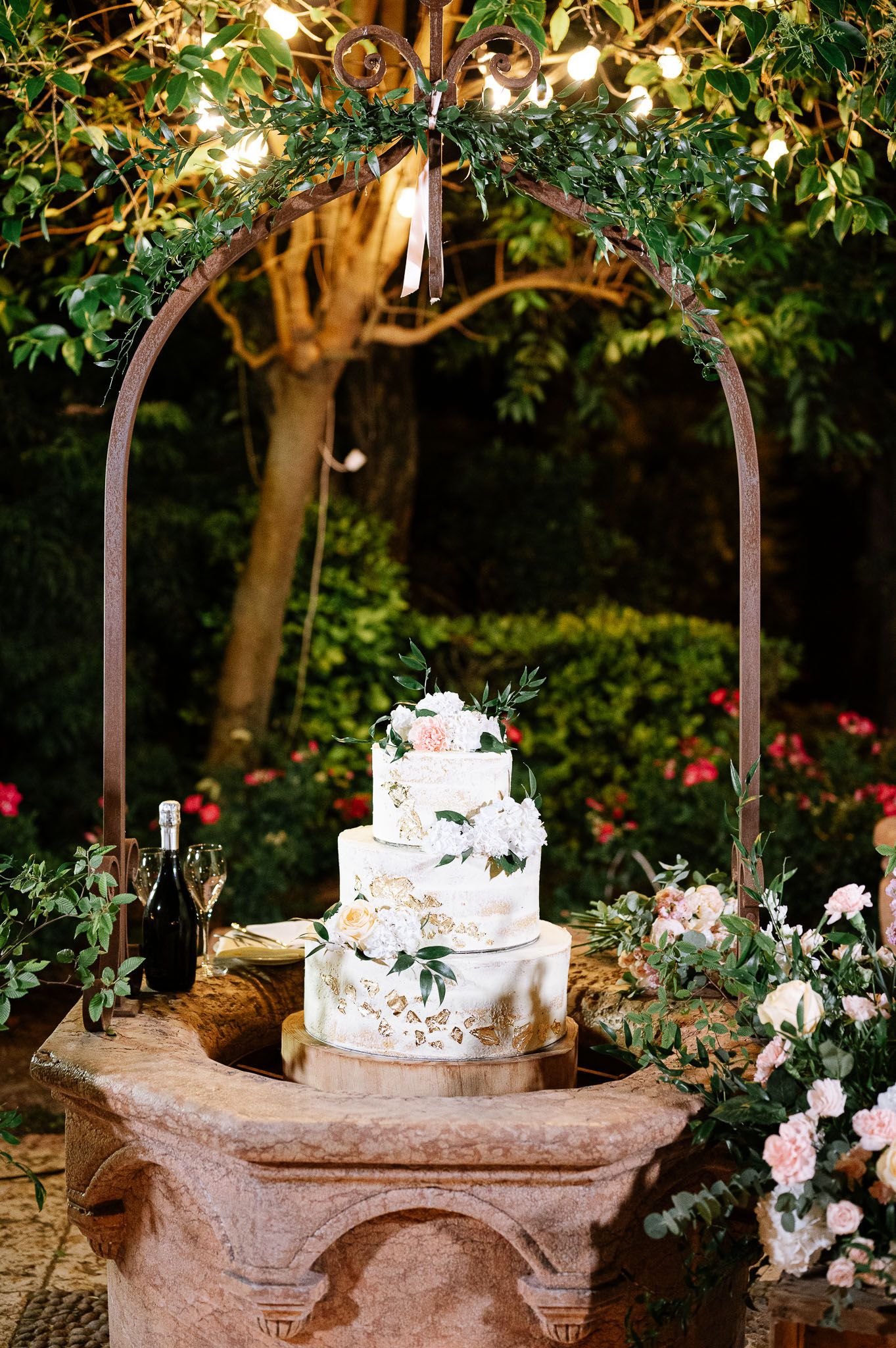 Wedding cake display under garden arch with twinkling lights and greenery in romantic outdoor setting.