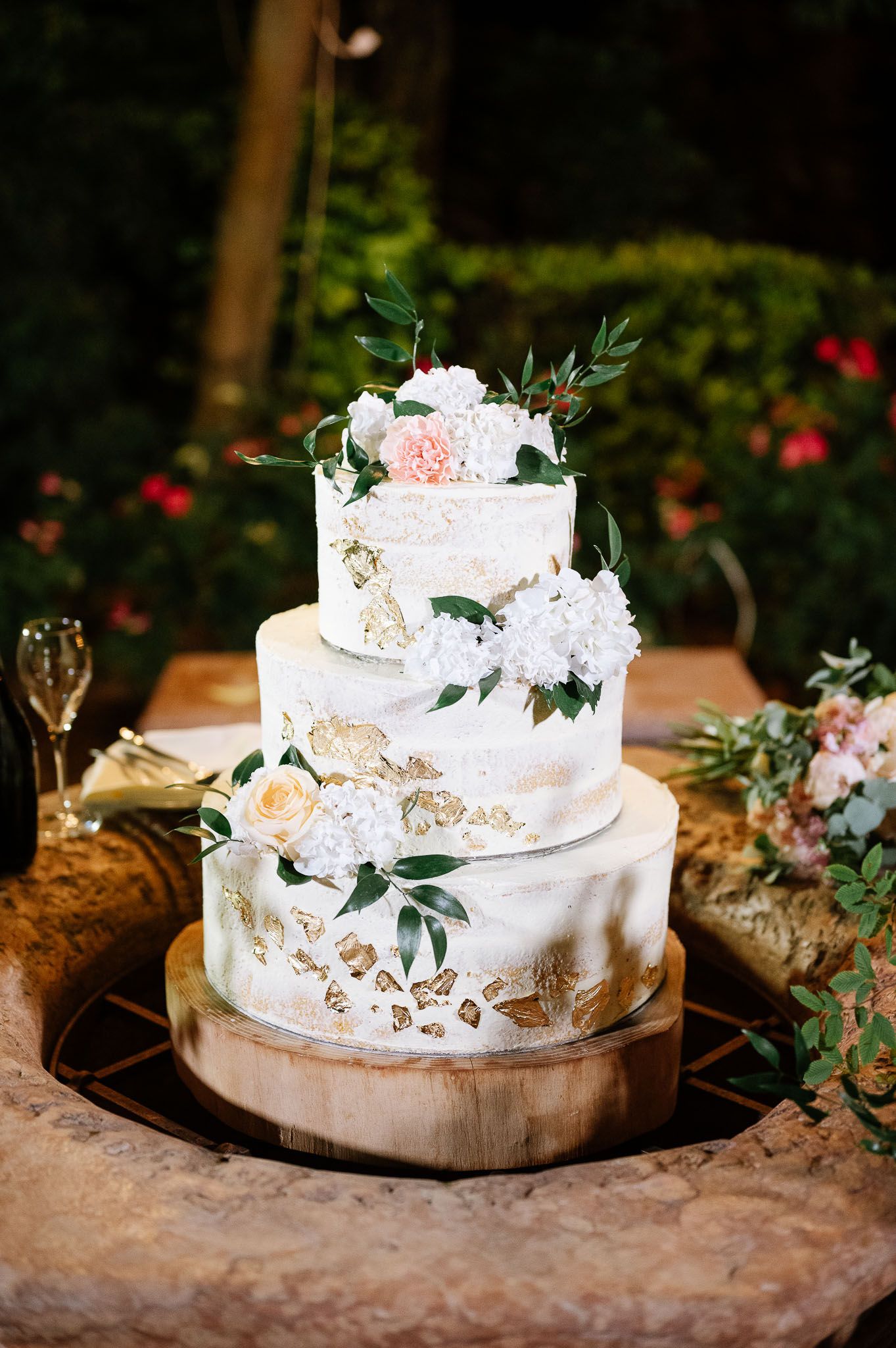 Four-tiered white wedding cake with gold accents and floral decorations displayed in garden setting.