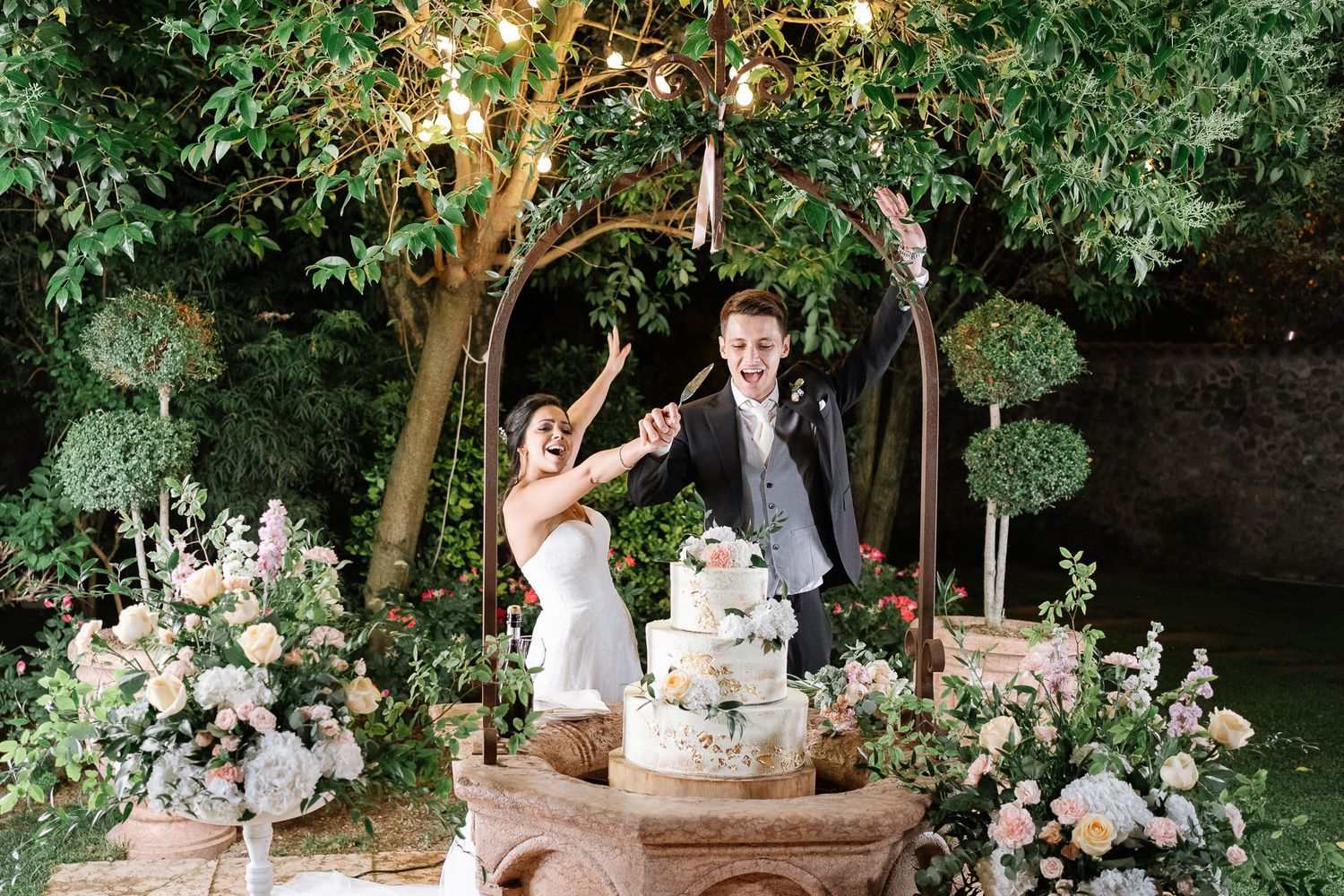 Celebratory moment at wedding cake cutting ceremony under illuminated garden arch with floral decorations.