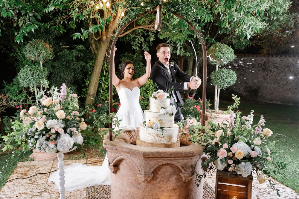 Wedding couple celebrates with raised arms during cake cutting ceremony in beautifully decorated garden setting.