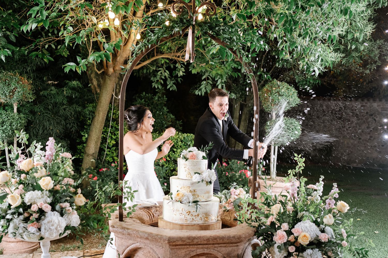 Wedding celebration in garden setting with ornate cake display and cascading floral arrangements.