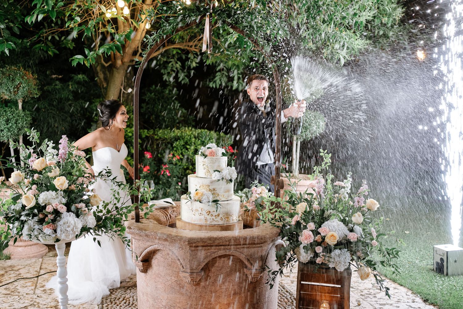 Festive wedding moment with water fountain spray adds dramatic effect to outdoor cake cutting ceremony.