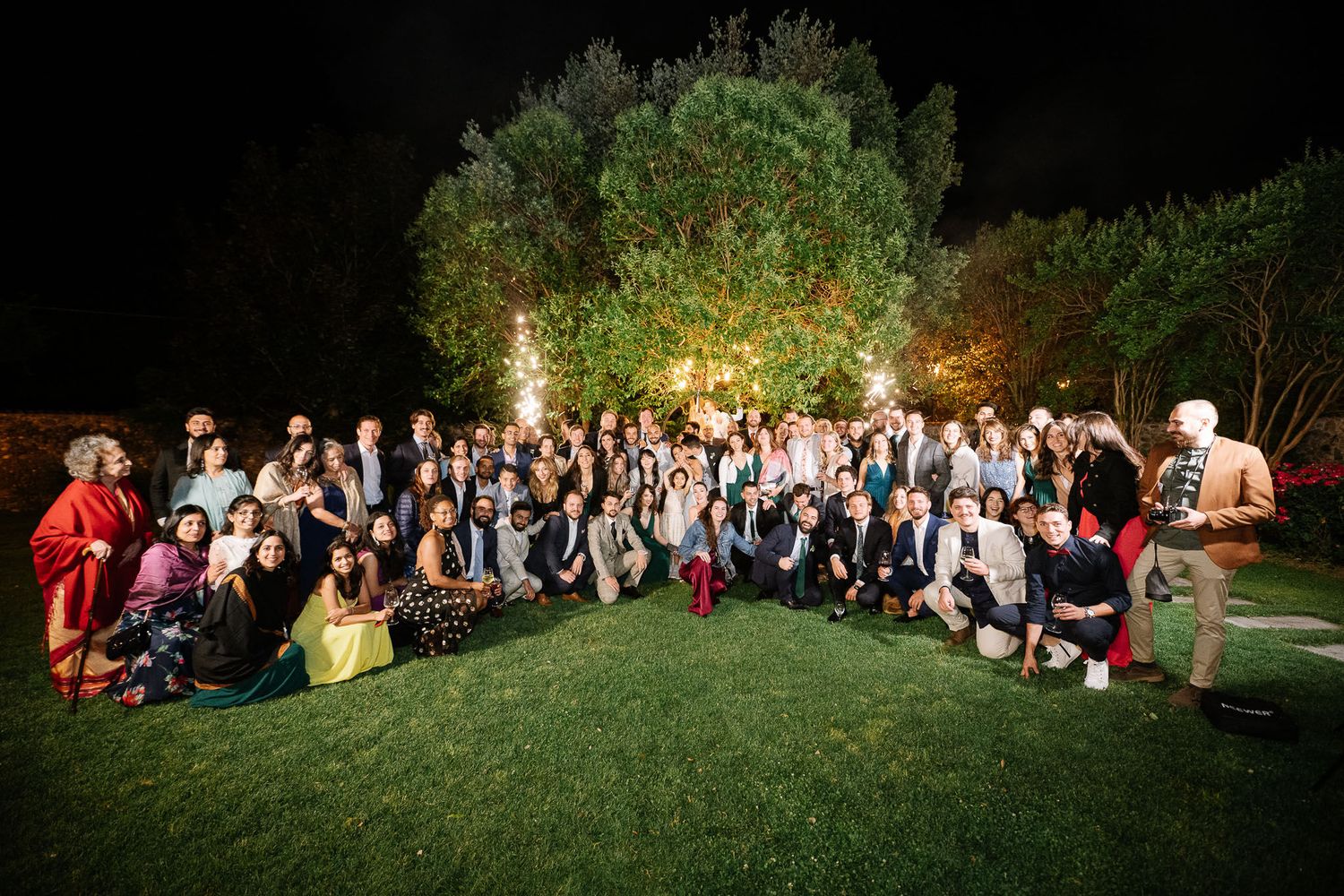 Large group wedding photo at night with illuminated trees creating magical outdoor ambiance.