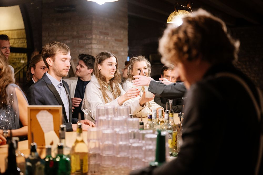 Wedding guests gather around a bar while watching drinks being prepared in a rustic venue.