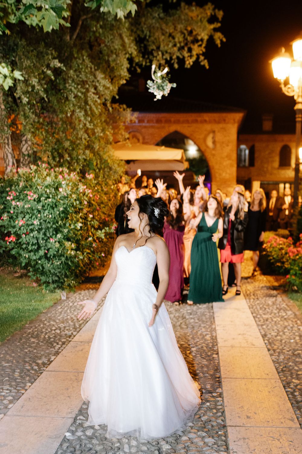 Bride in white gown walks along stone path while guests gather in background at evening celebration.