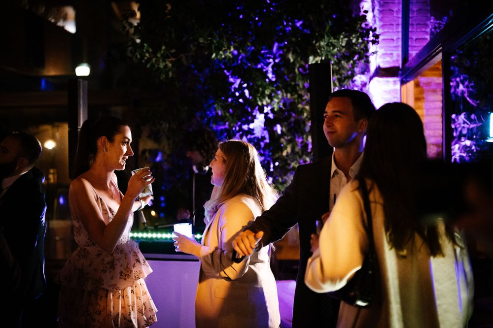 Party guests socializing and drinking under purple lighting at outdoor nighttime event.