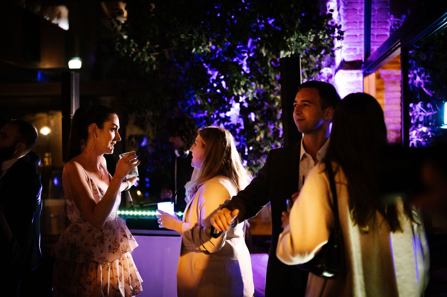 Party guests socializing and drinking under purple lighting at outdoor nighttime event.