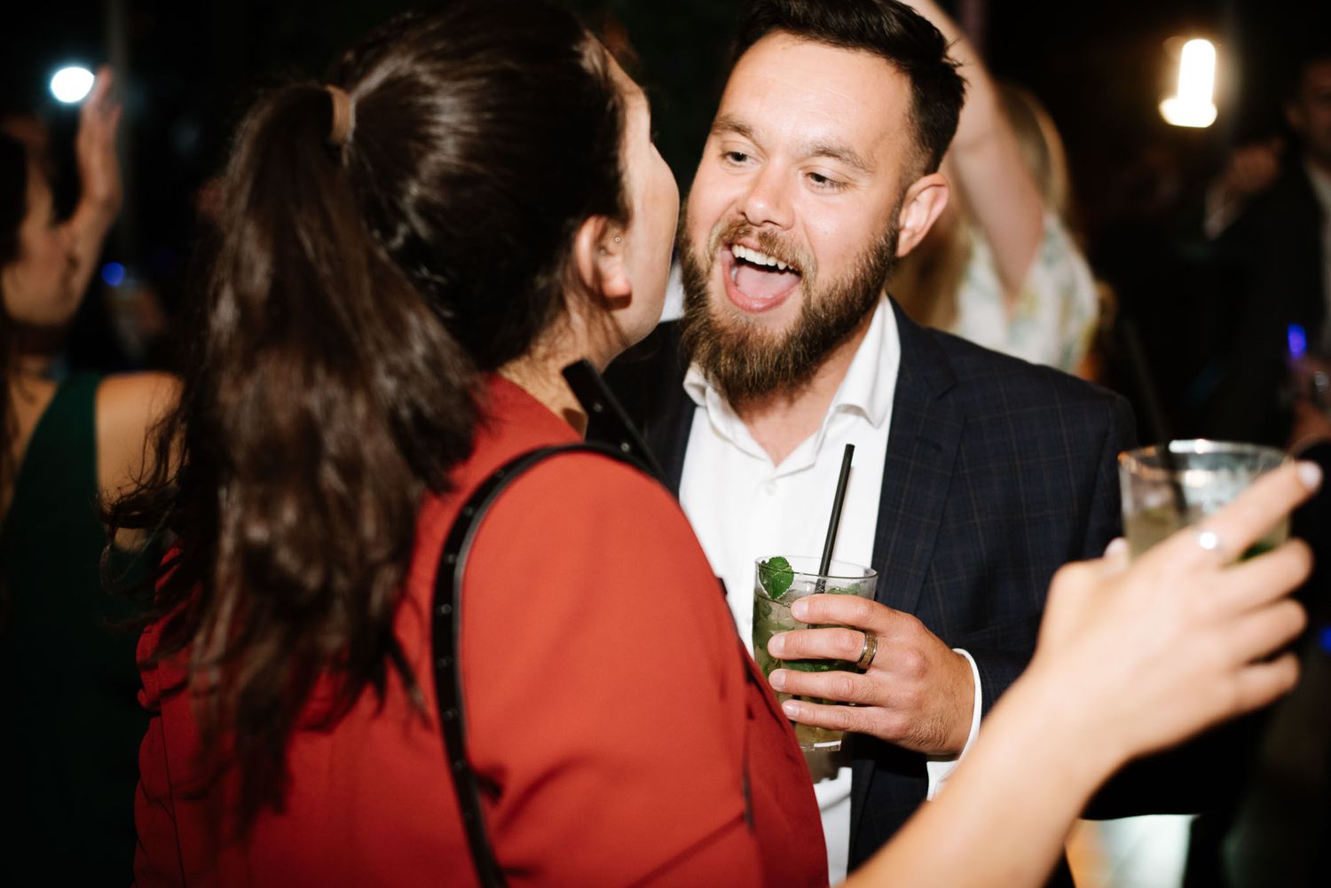 Two party guests share an animated conversation while holding drinks at a nighttime celebration.