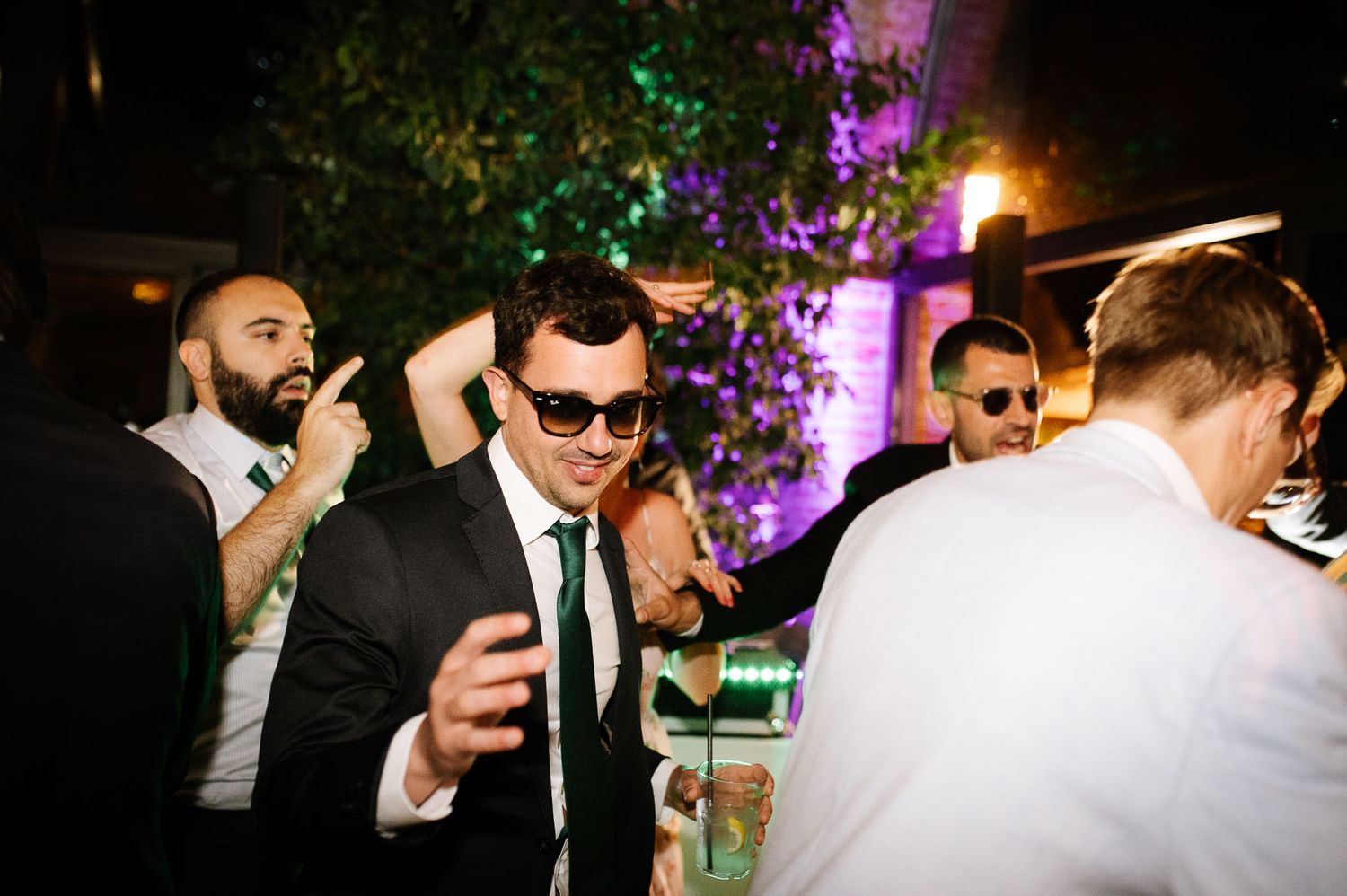 Stylishly dressed guests enjoying drinks and conversation at an evening party with purple lighting.