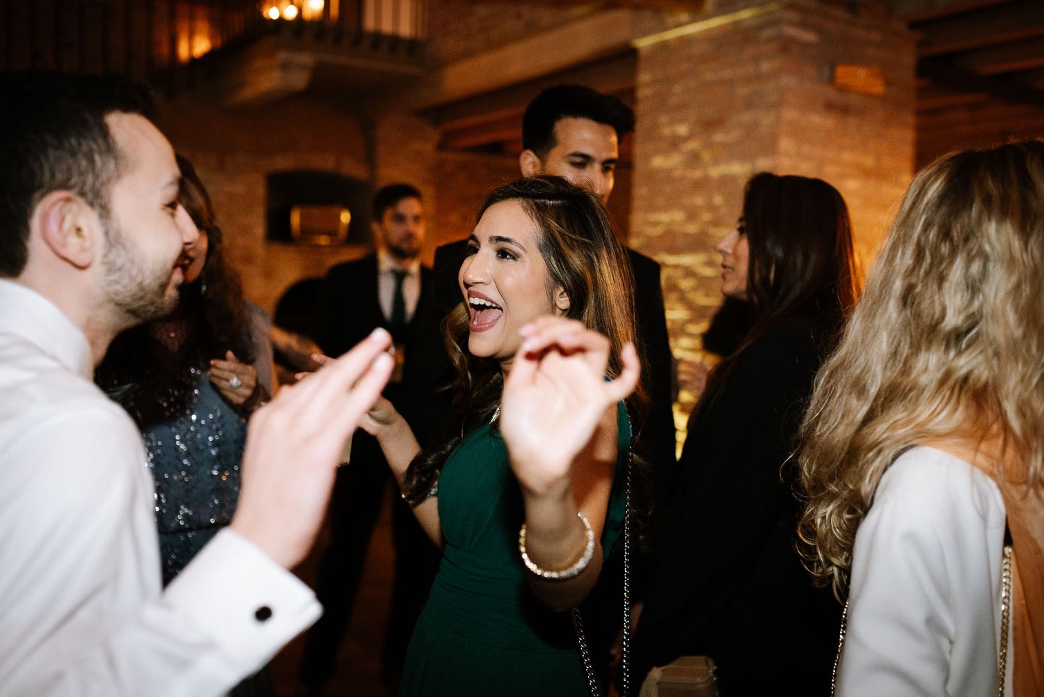 Wedding guests dancing and laughing energetically at a reception in a rustic brick venue.