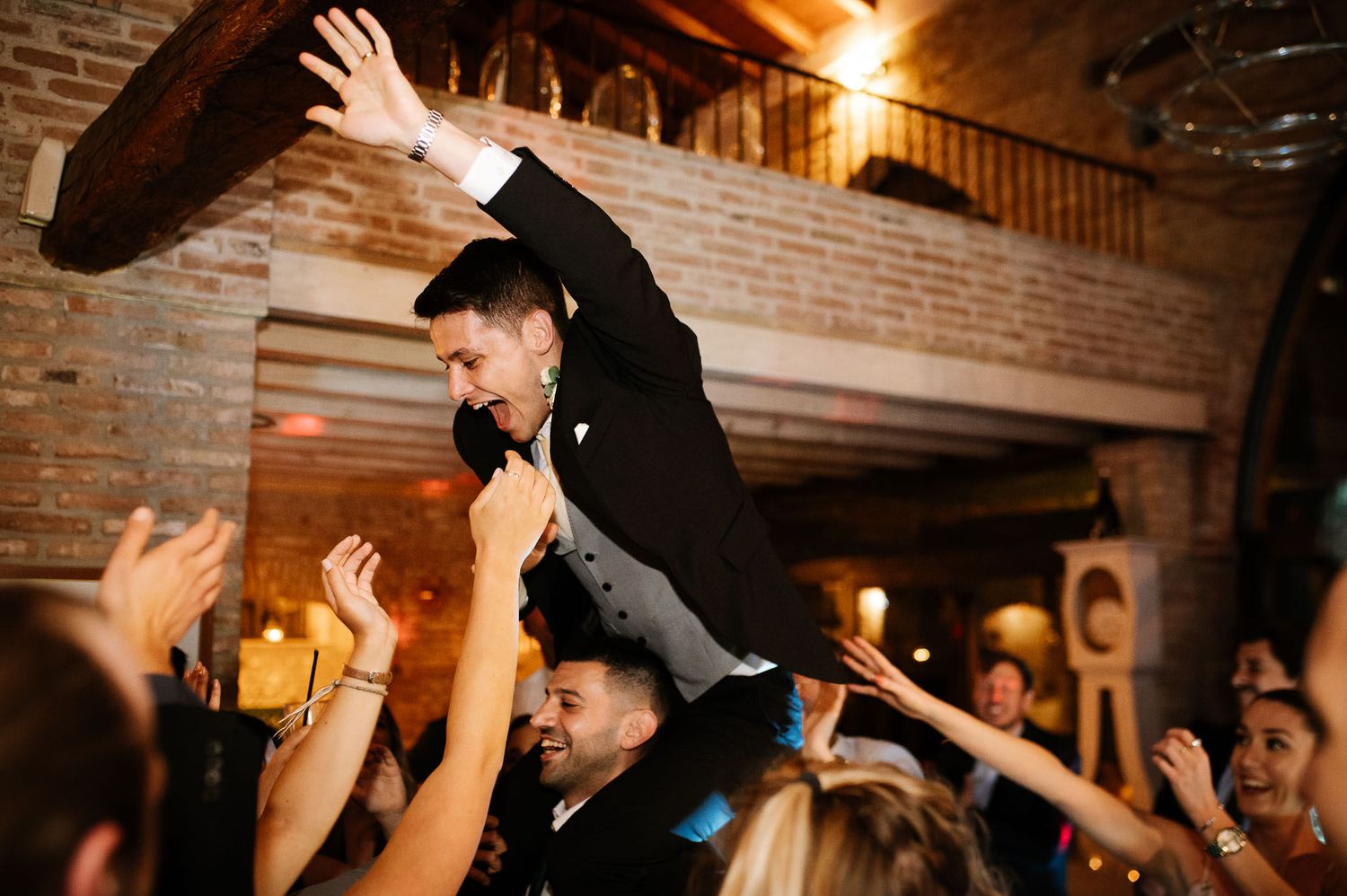 Guest in formal attire being lifted by crowd during celebratory moment at indoor venue.