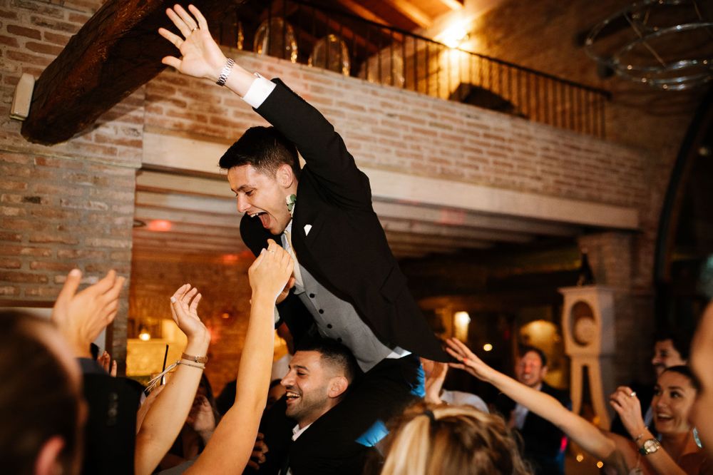 Guest in formal attire being lifted by crowd during celebratory moment at indoor venue.