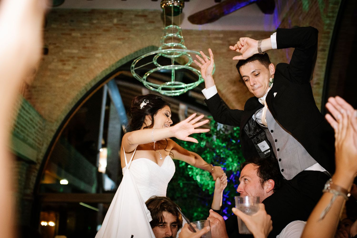 Reception guests dancing and celebrating beneath elegant chandelier in rustic venue.
