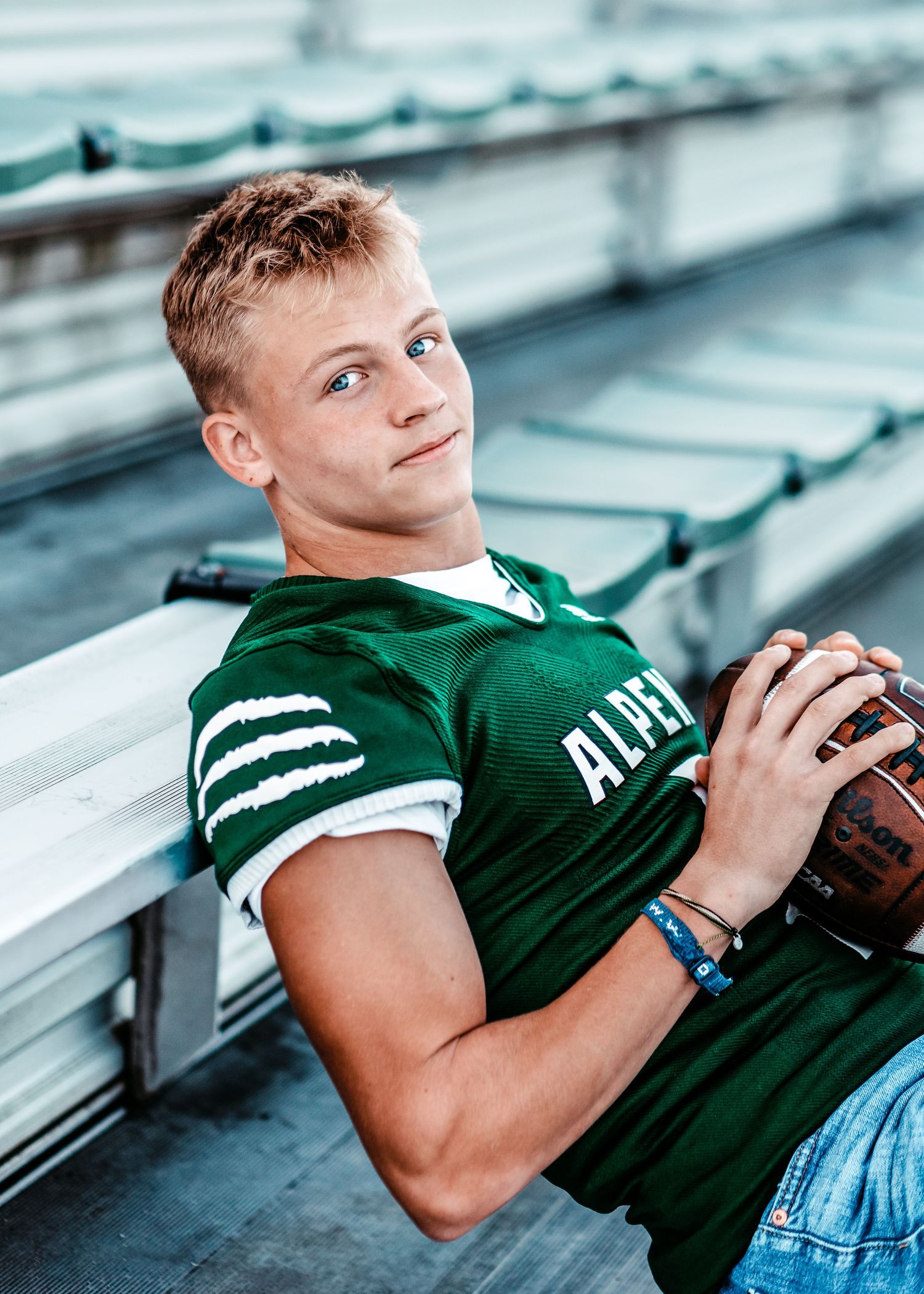 Student athlete in Alpena football jersey poses with football in bleachers.