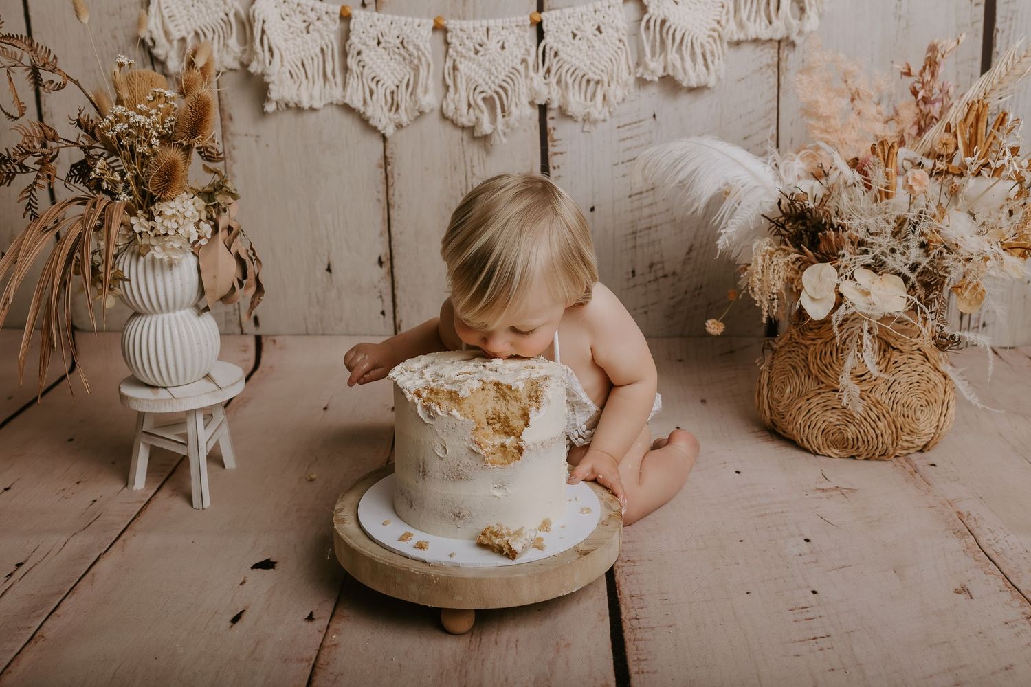 Rustic cake smash photography setup with a white cake and neutral dried flowers against a weathered wood backdrop.