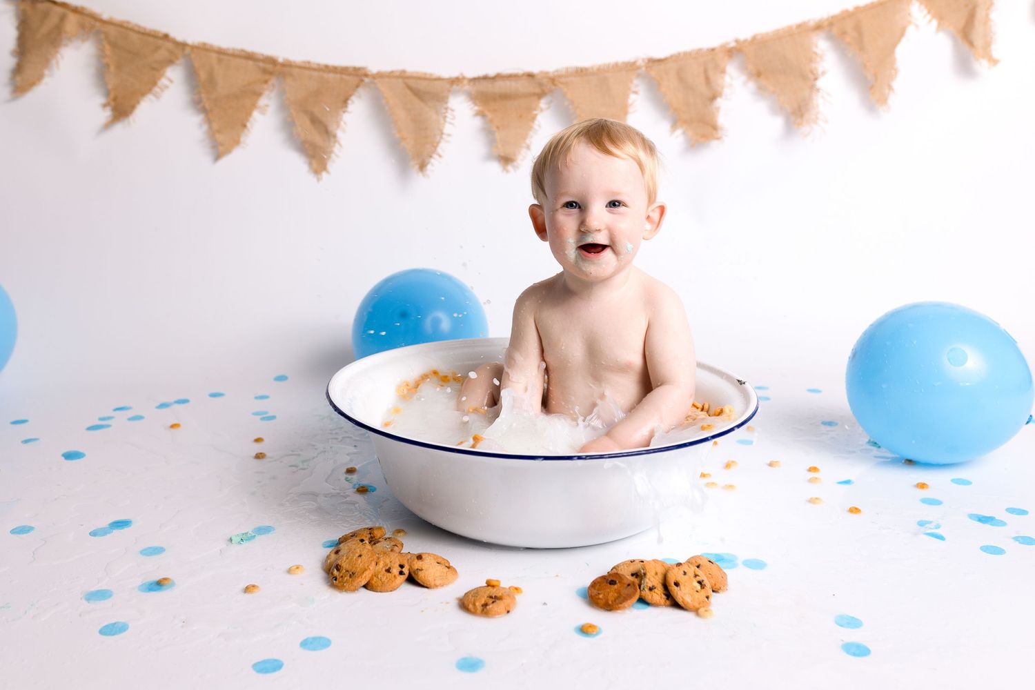 Baby sitting in white basin with scattered cookies and blue balloons against burlap banner.
