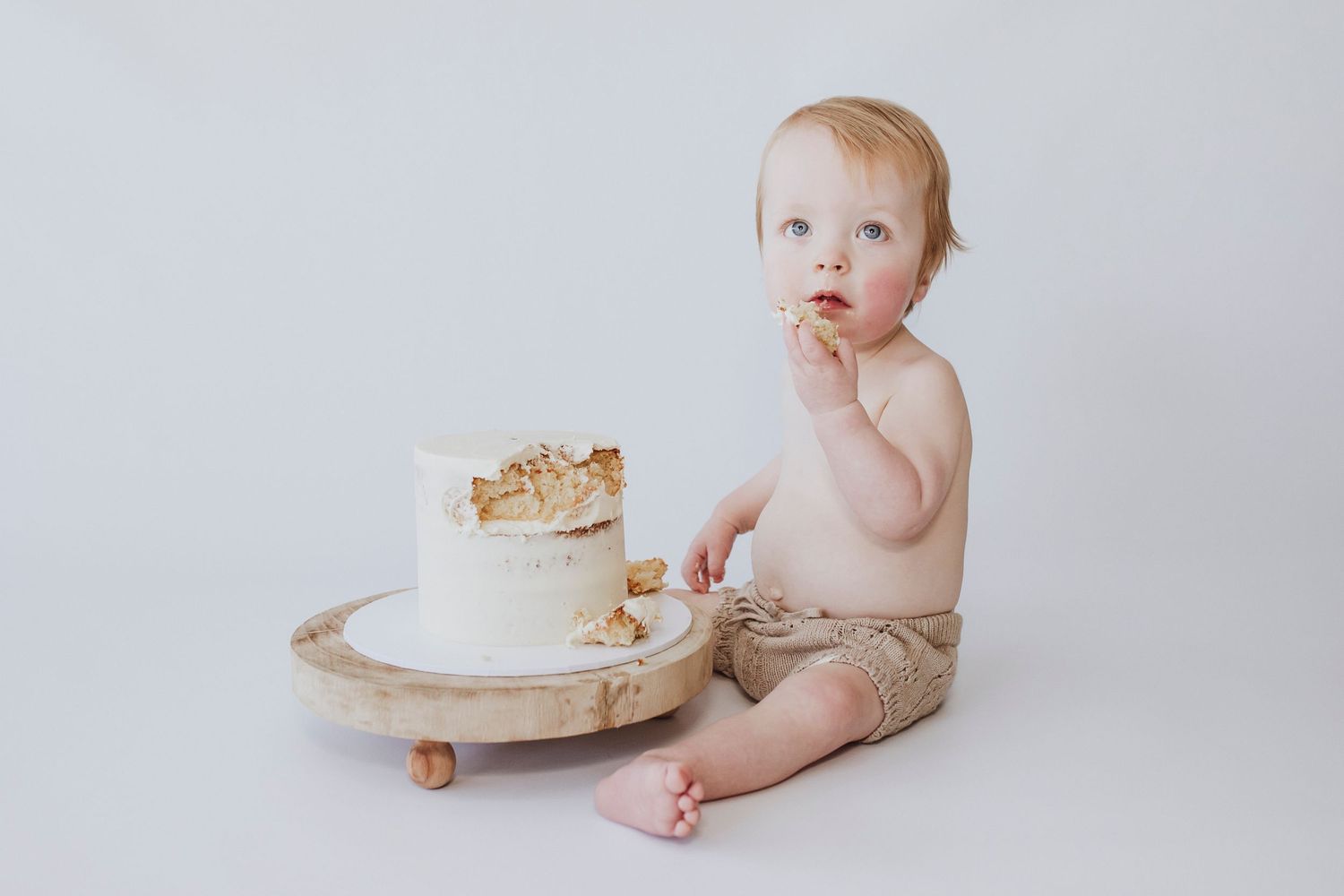 Baby exploring white birthday cake on wooden plate against minimal white background.