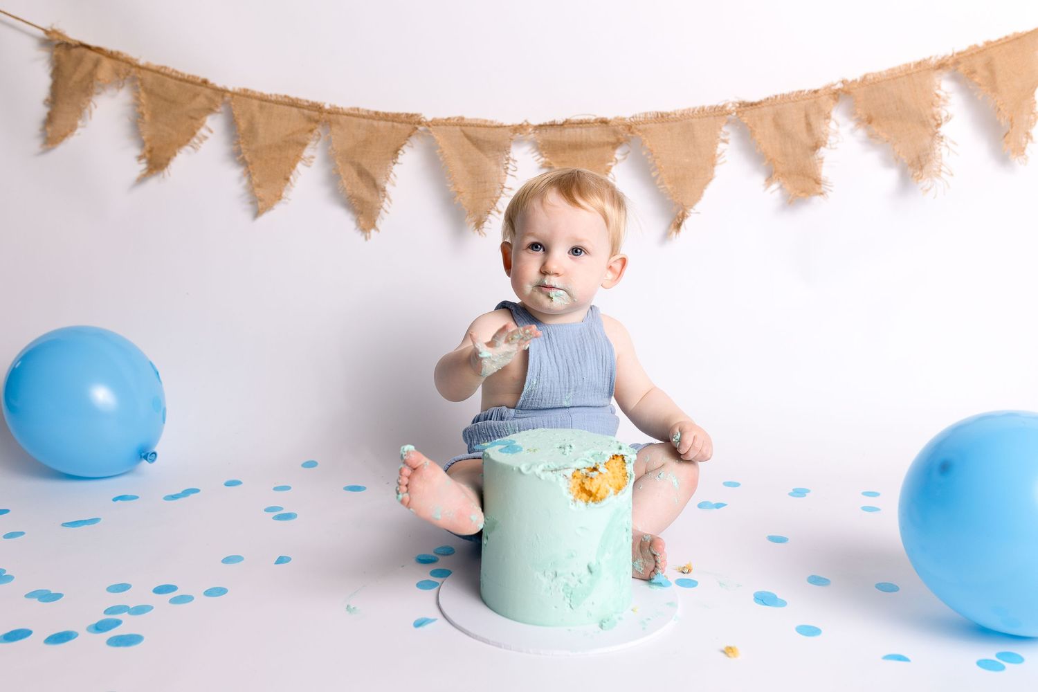 Baby in blue overalls sitting on mint green prop surrounded by blue balloons and confetti.