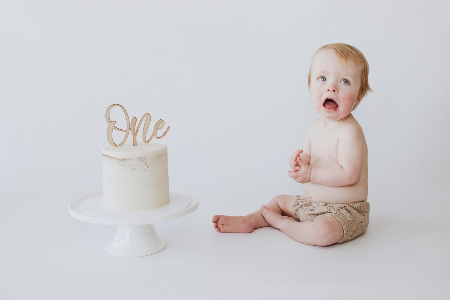 Baby sitting beside white cake with 'One' topper on minimalist white backdrop.