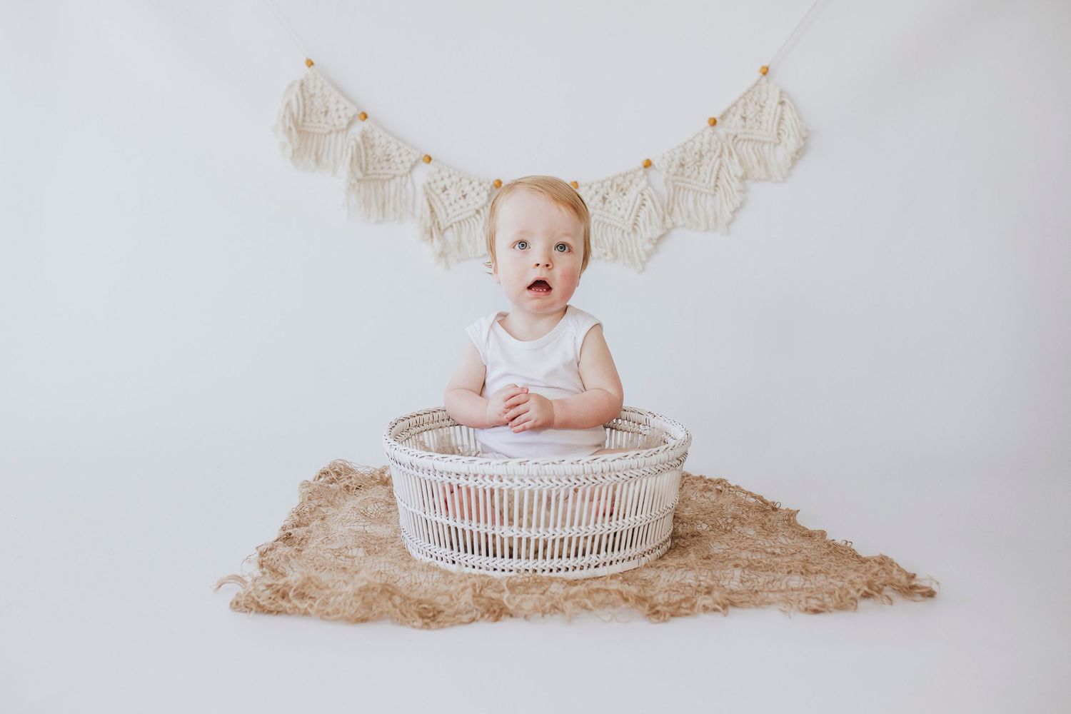 Baby sitting in white wicker basket with burlap bunting banner overhead.