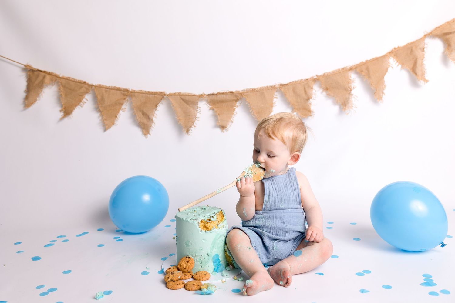 Baby in blue outfit enjoying cookies during cake smash photoshoot with blue balloons.