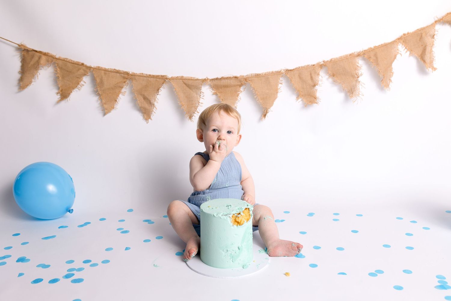 Baby sitting on a mint green cake smash prop with blue balloons and burlap bunting banner backdrop.