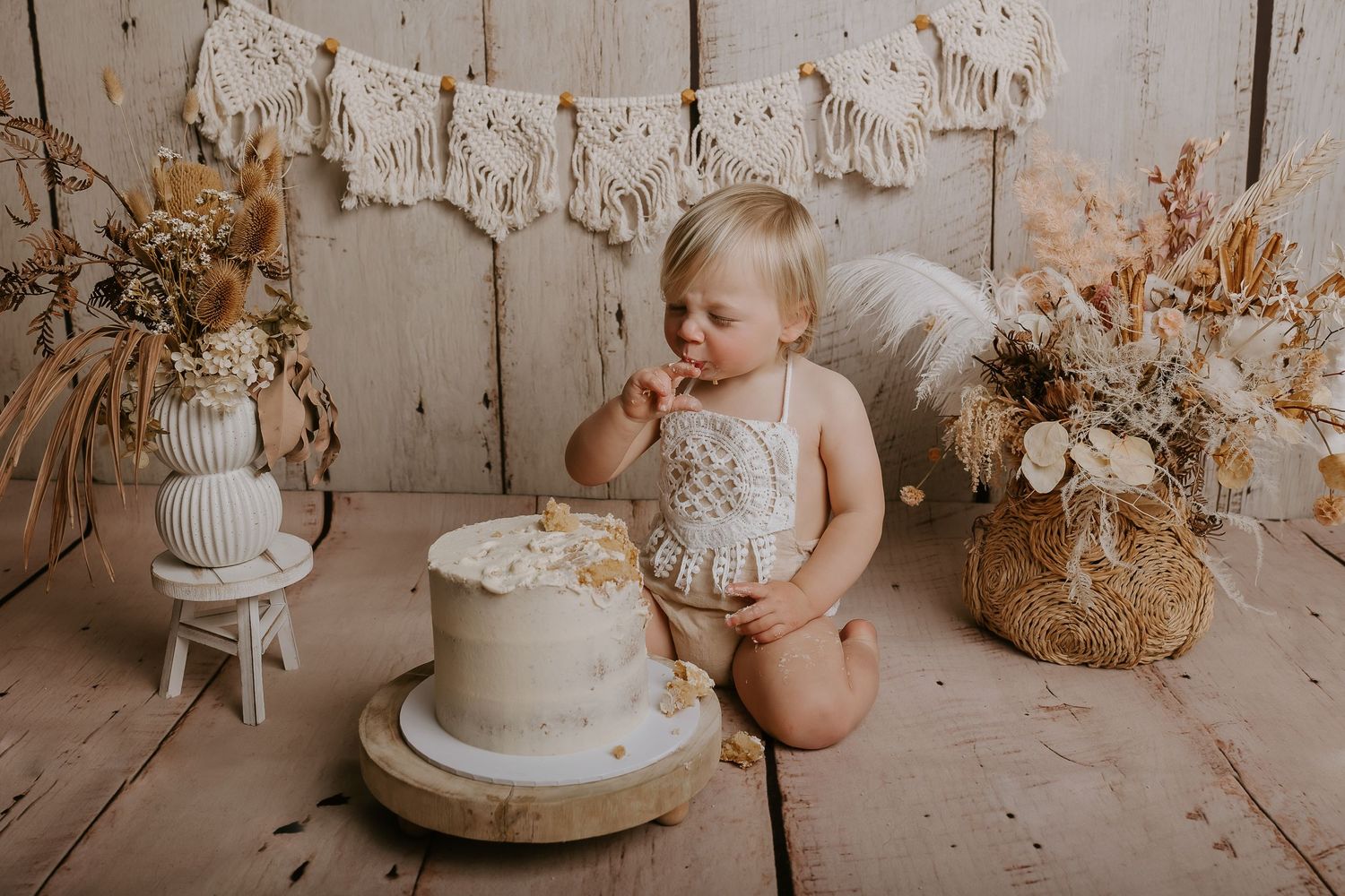 A white birthday cake with neutral decorations sits on a rustic wooden floor next to dried floral arrangements.