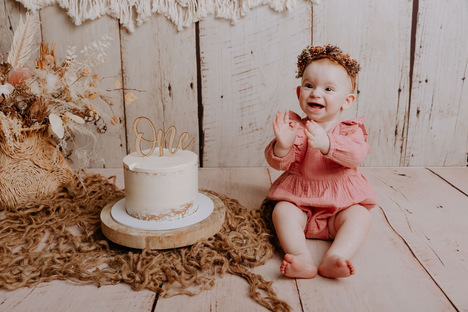 Baby in pink romper sitting next to white cake with macrame banner decoration.