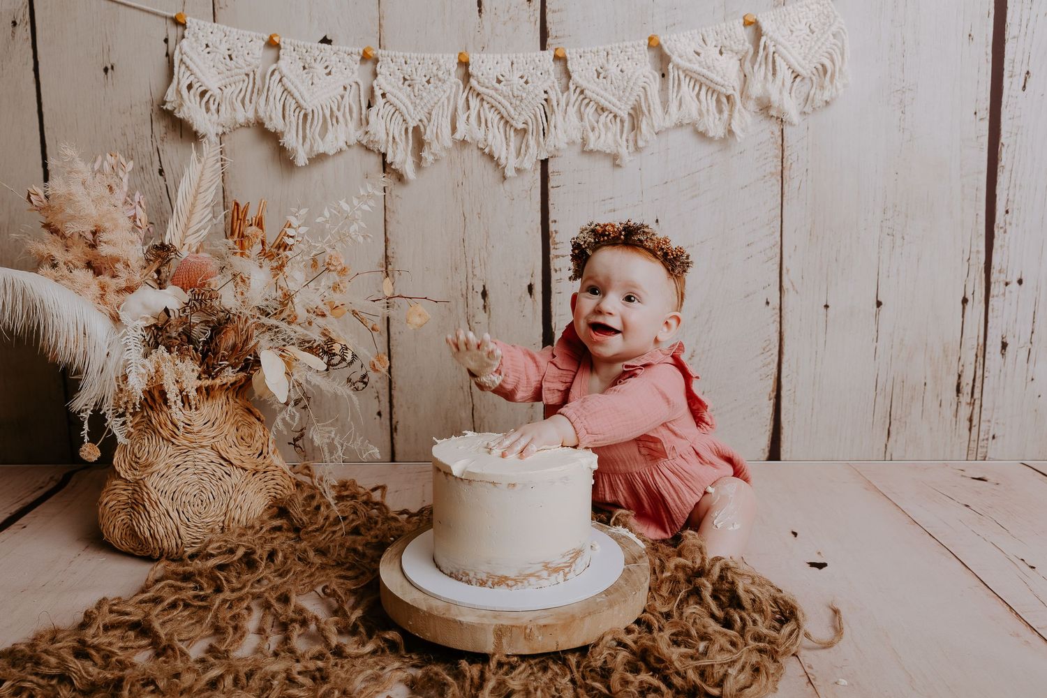 Baby in pink outfit laughing next to white cake with rustic decor and macrame banner.