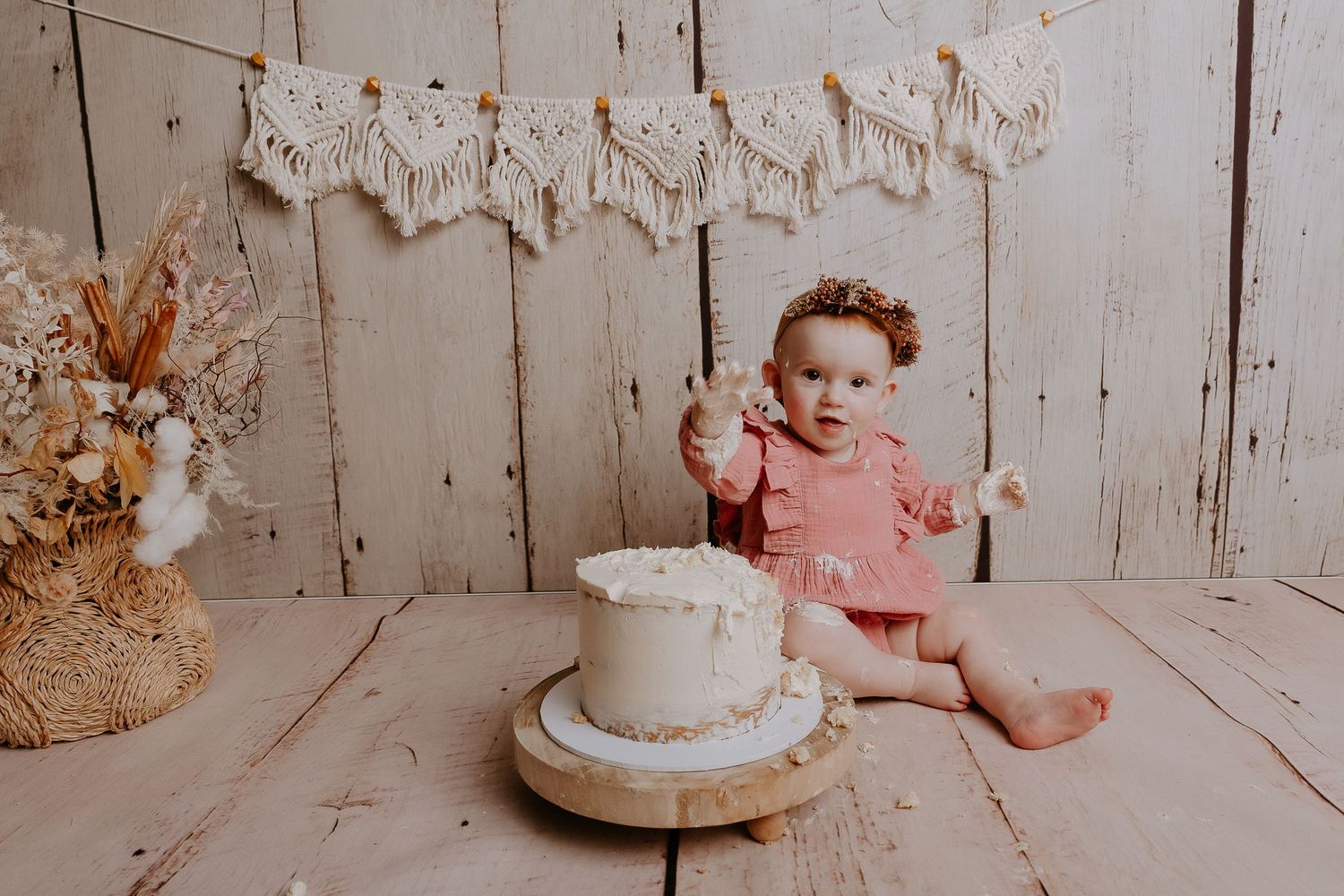 Baby sitting on wooden floor with decorative macrame banner and white cake.