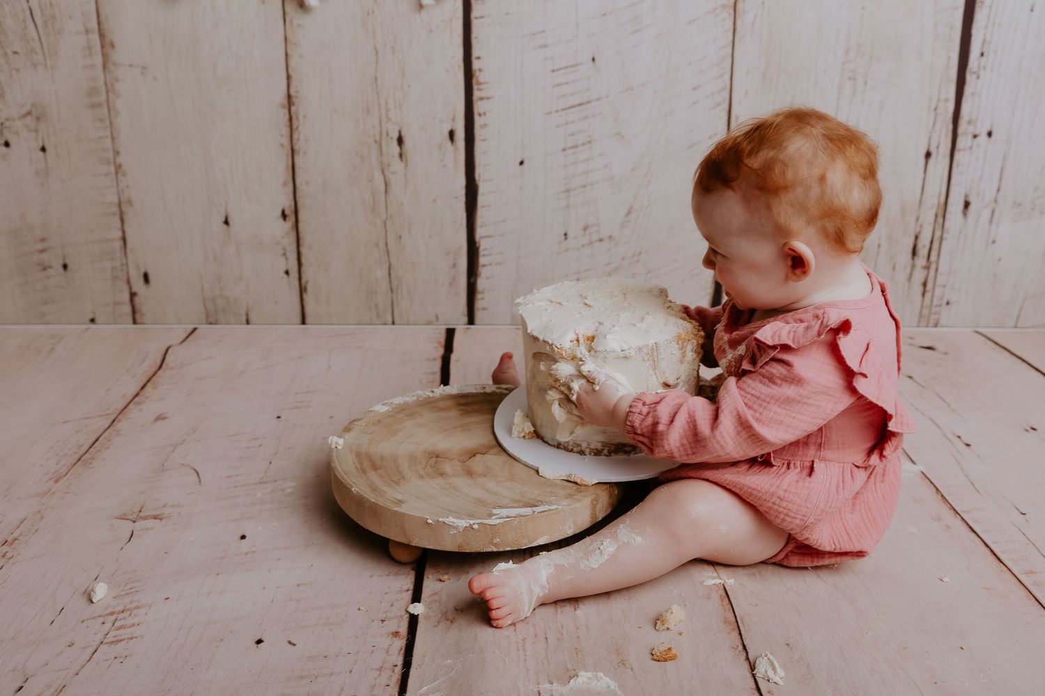 Baby in pink outfit reaching toward white cake on wooden floor.