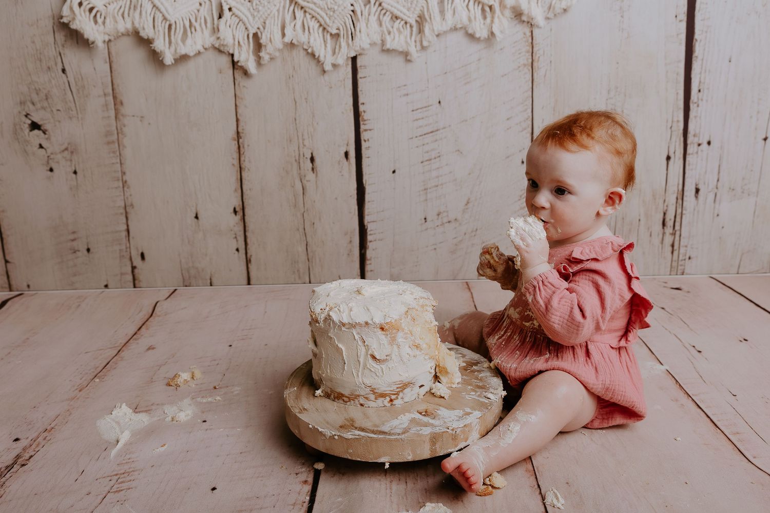 Baby in pink outfit next to white cake with macrame wall decoration.