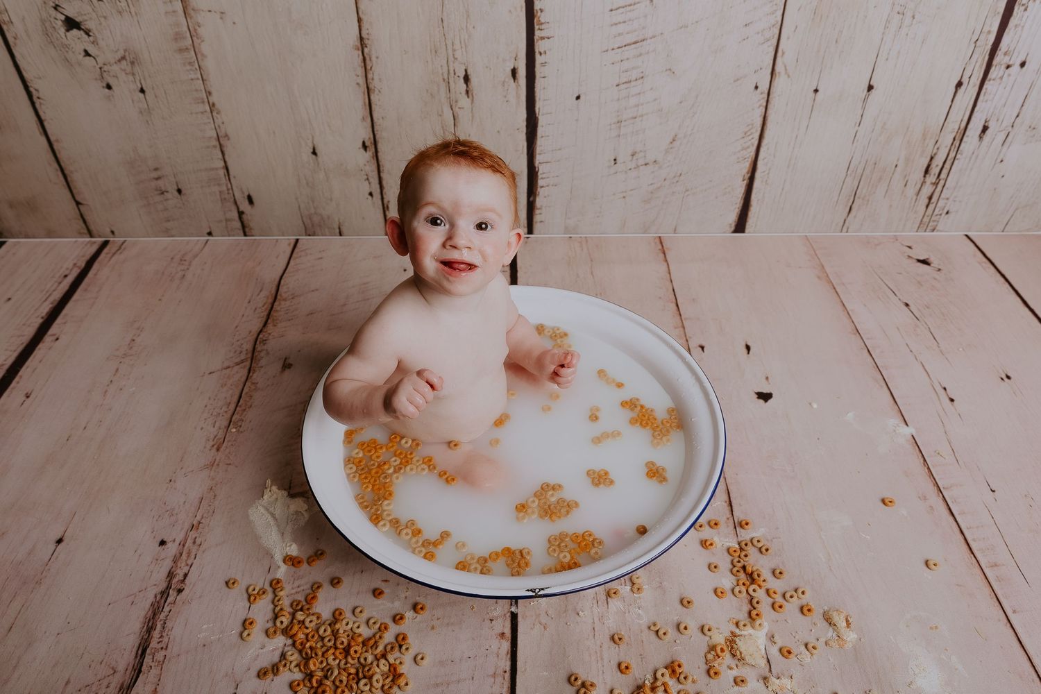 Baby having fun with cereal scattered around white bowl on wooden floor.