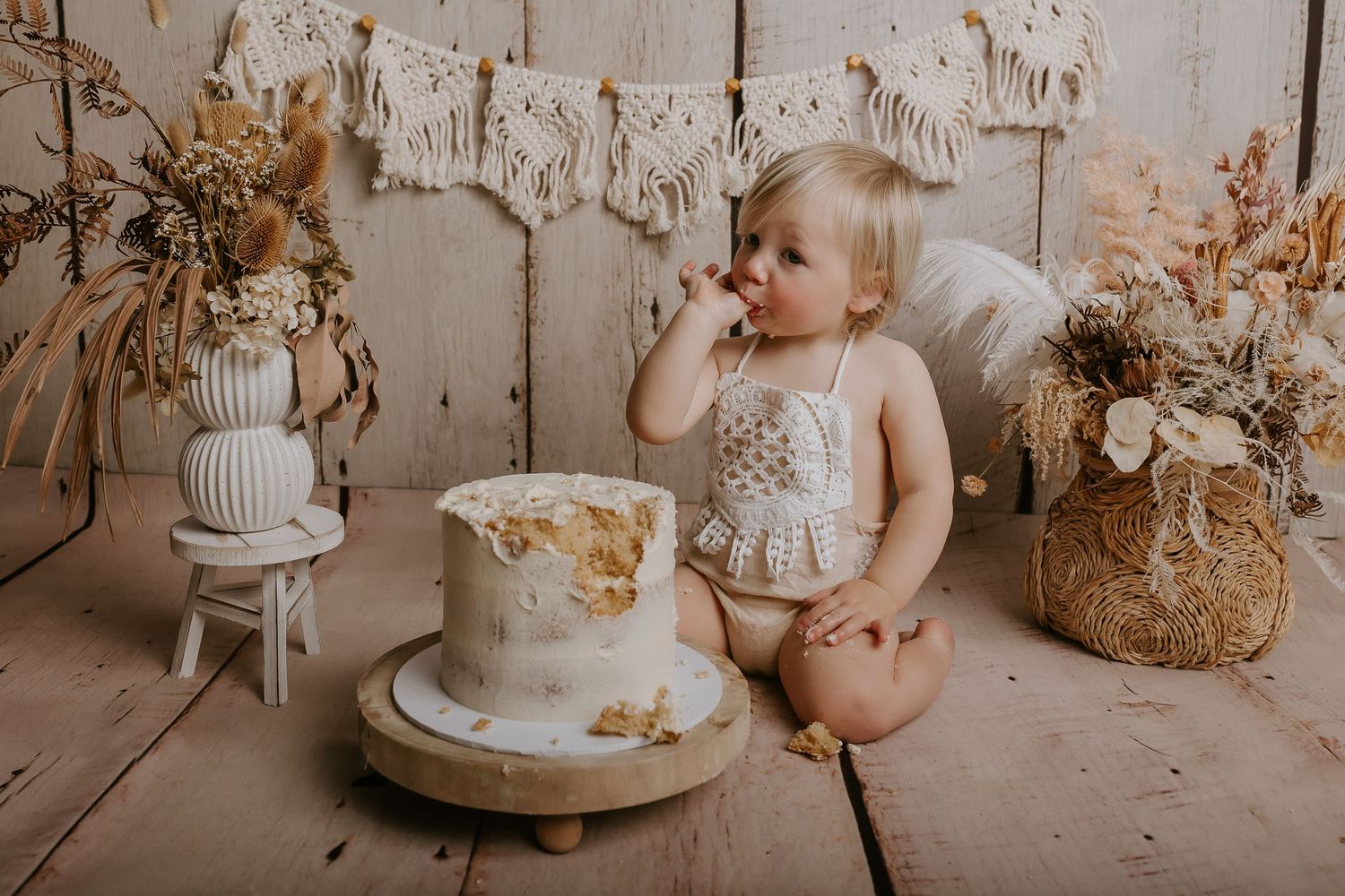 Minimalist cake smash scene with white frosted cake and beige dried flowers against whitewashed wood walls.