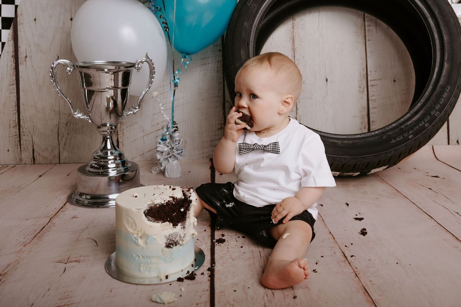 Baby wearing white onesie sits among vintage props and black and white checkered banner decorations.
