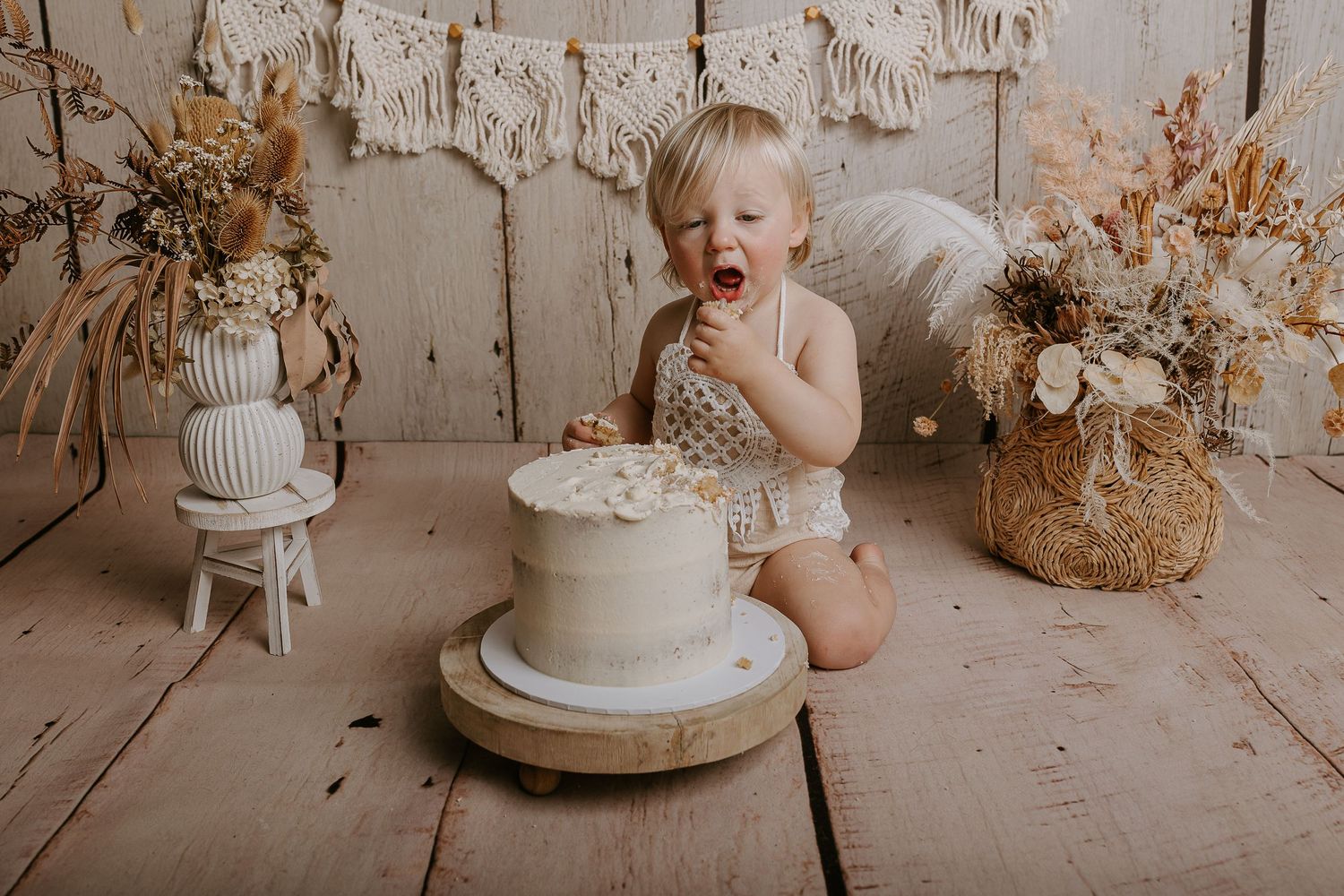 Baby in white outfit reaching for cake during rustic themed photo session.