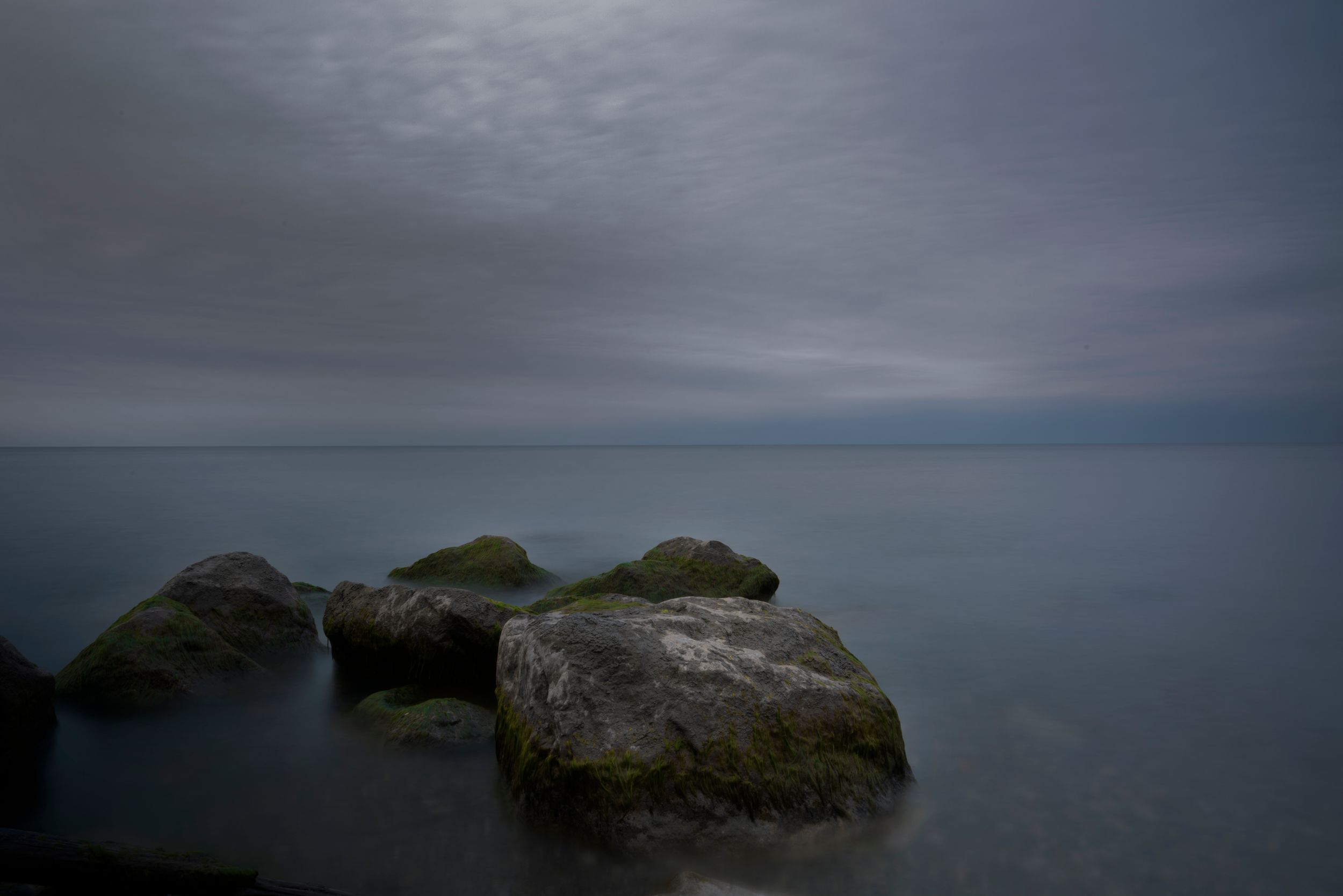 Mossy rocks emerge from calm water under moody gray skies in low light seascape.