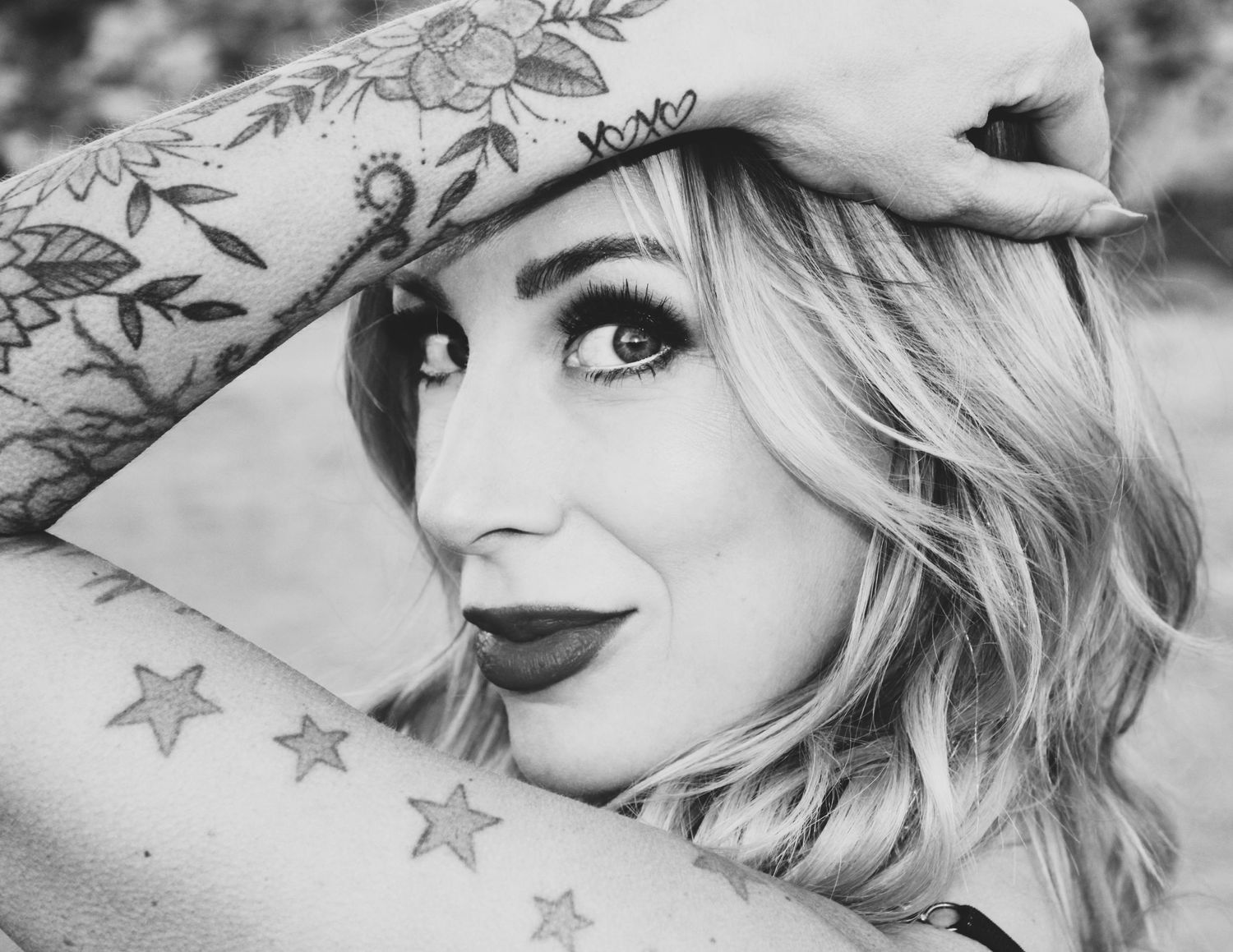 Black and white portrait showing a person with wavy blonde hair, dark lipstick and arm tattoos posing with hand raised.
