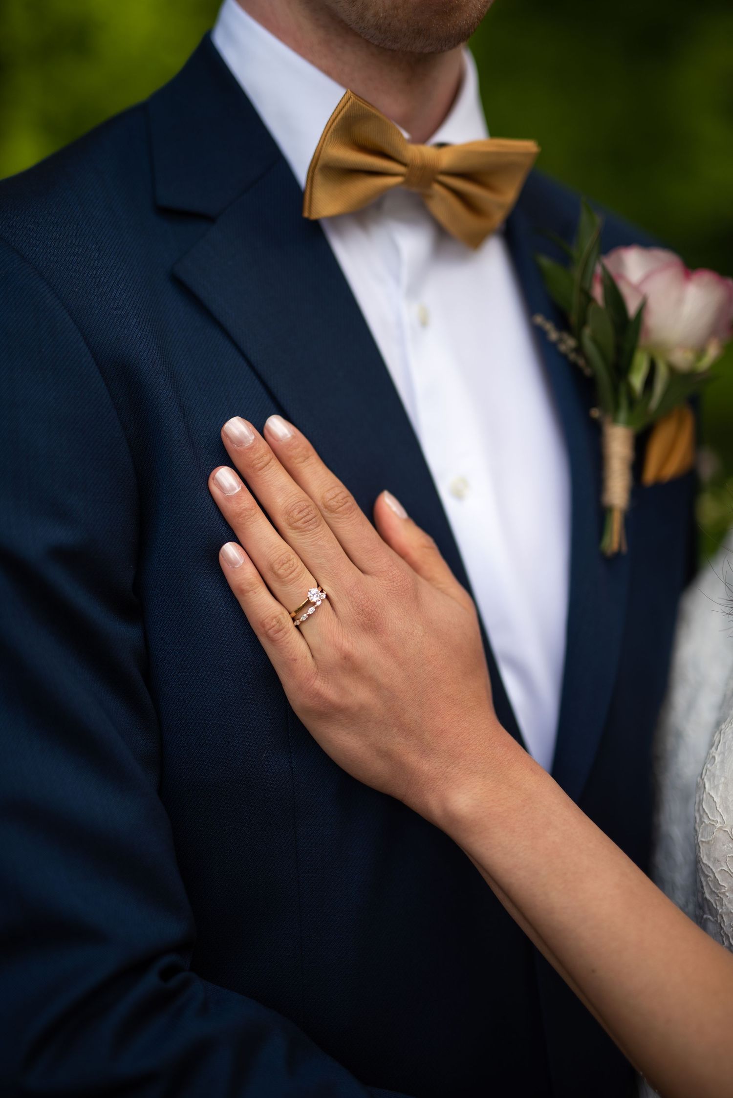 Bride places her hand on grooms navy suit jacket, showing her wedding ring beside a gold bow tie boutonniere.