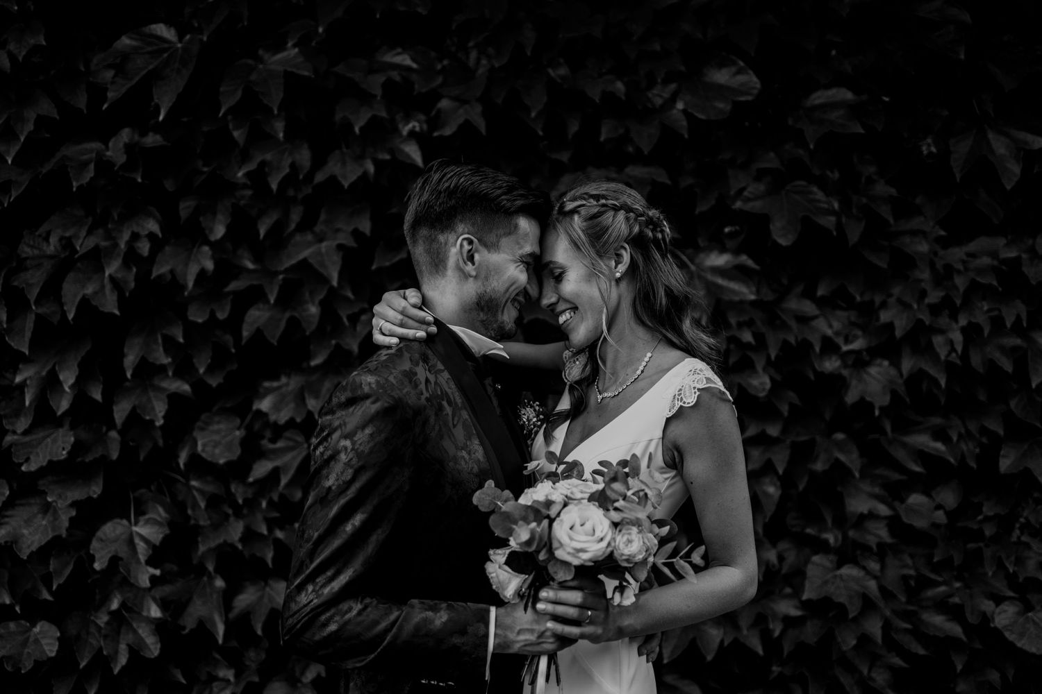Black and white romantic portrait of bride and groom embracing in front of lush ivy wall.