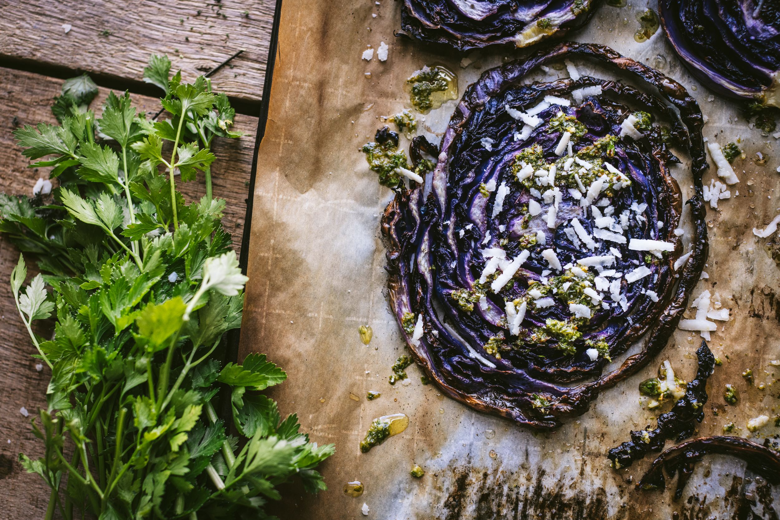 Grilled purple cabbage steaks with pesto and cheese on parchment paper beside fresh herbs.