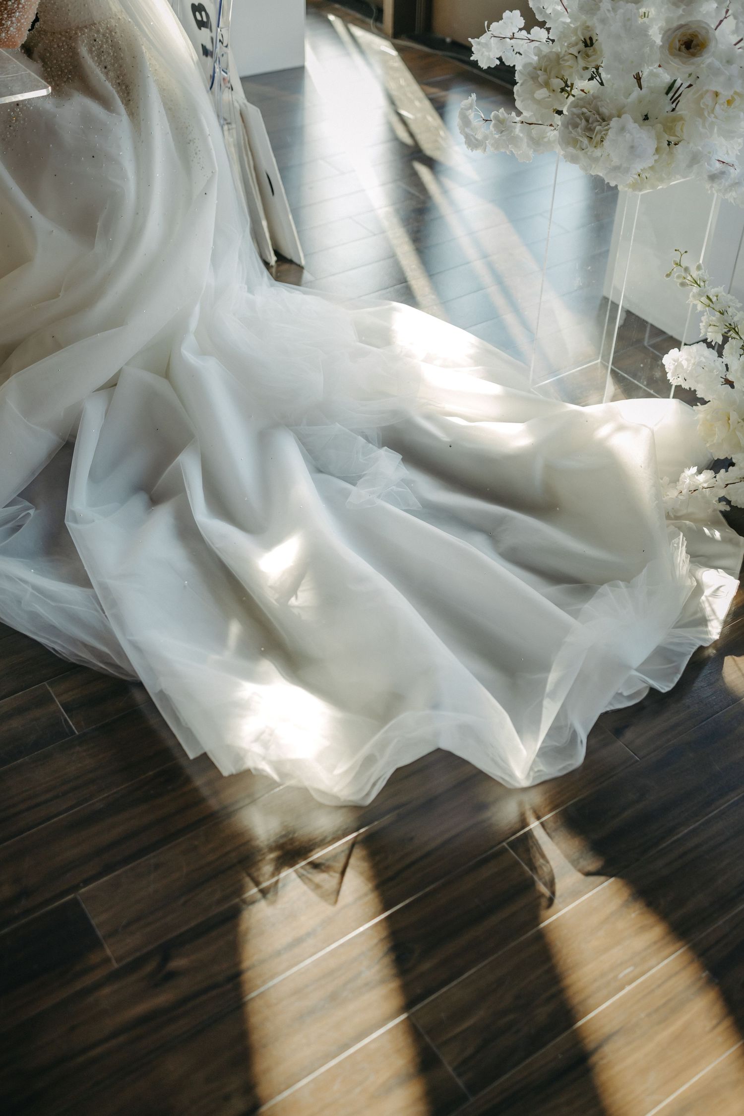 White flowing wedding dress fabric draped elegantly on a hardwood floor with sunlight streaming across.