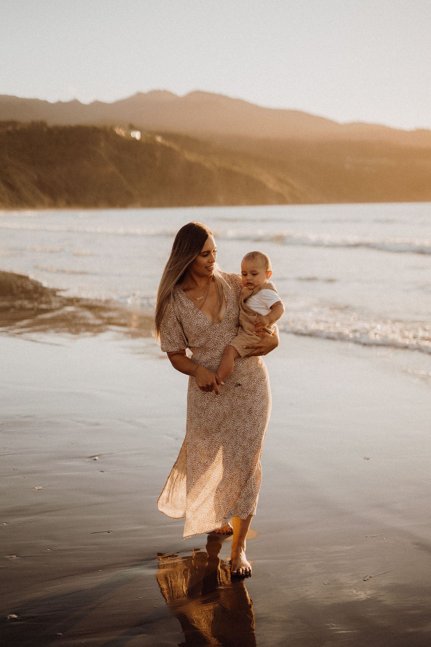 Mother and baby walking along beach shore at sunset in flowing dresses.